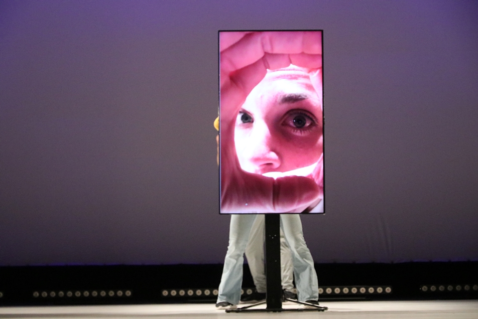 Personen stehen hinter einem Bildschirm, auf dem Bildschirm ist in Großformat gezeigt, wie eine Person durch die zu einer Röhre geformten Hand guckt