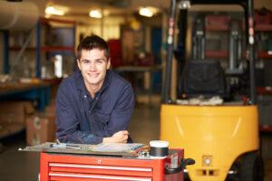 Symbolbild, in einer Werkhalle stützt sich ein junger Mann in Arbeitskleidung auf einen Werkzeugcontainer; Gabelstapler im Hintergrund