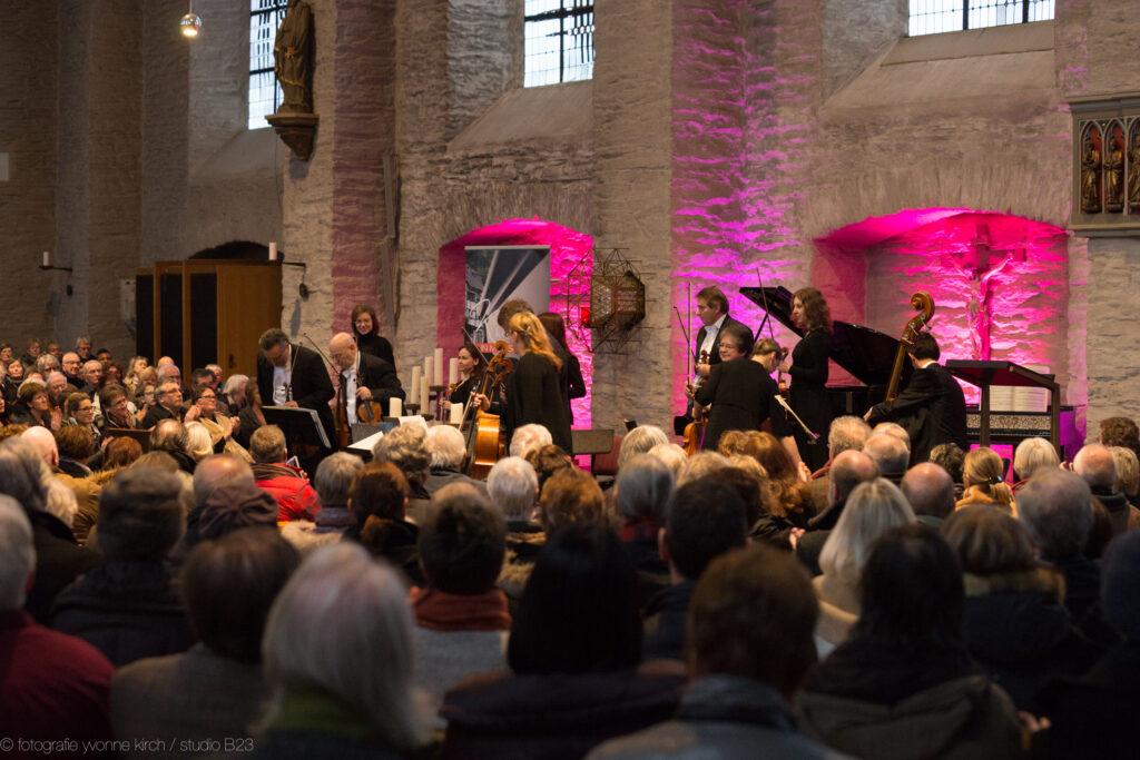 Konzert ine iner Kirche, Publikum von hinten im Bild, auf der Bühne verschiedene Streicher pink ausgeleuchtet